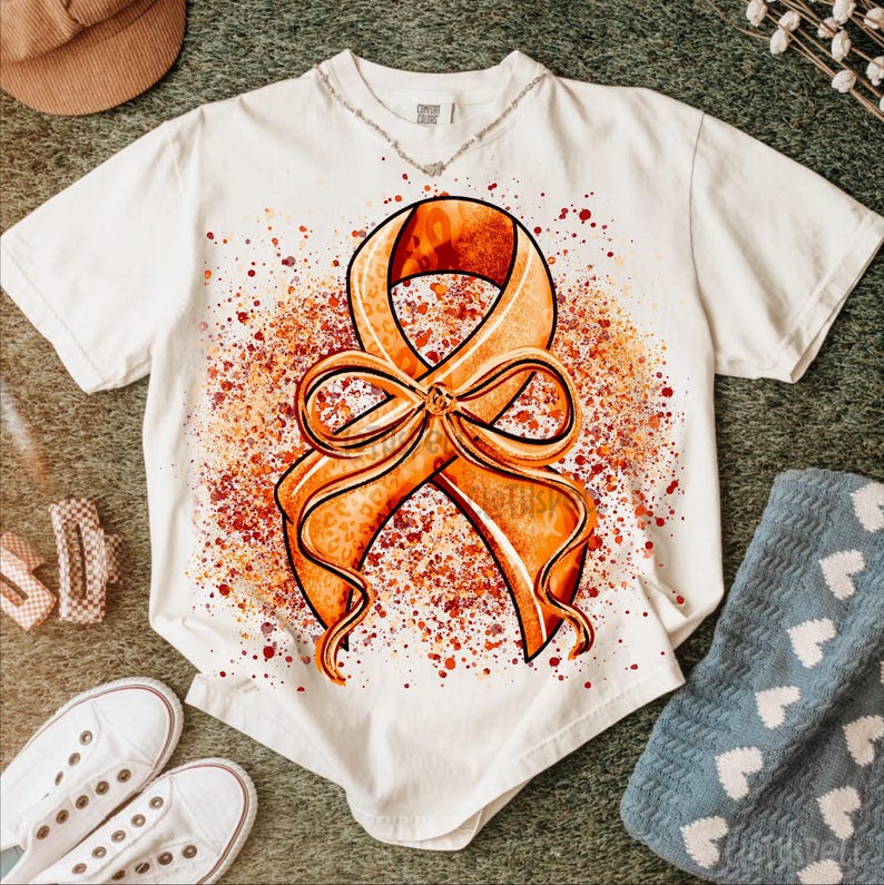 May include: White t-shirt featuring a large orange ribbon design with a leopard print pattern and a bow. The ribbon is set against a backdrop of orange and red paint splatters. The shirt is surrounded by accessories.