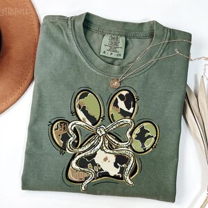 K&ouml;nnte beinhalten: Olivgr&uuml;nes T-Shirt mit einem Pfotenabdruck-Design. Der Pfotenabdruck hat eine Schleife und Tarn- und Kuhmuster. Eine goldene Halskette mit Anh&auml;nger befindet sich auf dem T-Shirt. Das T-Shirt ist mit "Comfort Colors" gekennzeichnet.
