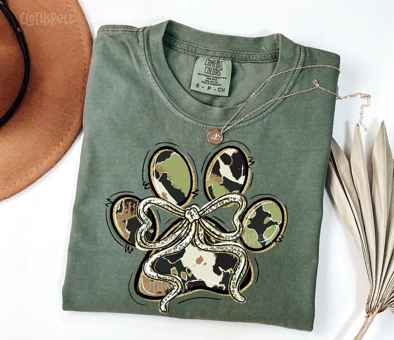 K&ouml;nnte beinhalten: Olivgr&uuml;nes T-Shirt mit einem Pfotenabdruck-Design. Der Pfotenabdruck hat eine Schleife und Tarn- und Kuhmuster. Eine goldene Halskette mit Anh&auml;nger befindet sich auf dem T-Shirt. Das T-Shirt ist mit "Comfort Colors" gekennzeichnet.