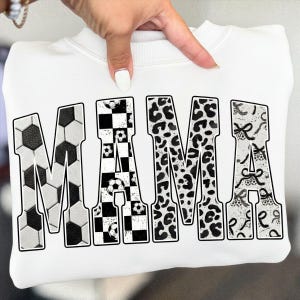 May include: White sweatshirt with the word "MAMA" in large, bold letters. The letters are decorated with soccer ball patterns, black and white checkered patterns, leopard print, and a ribbon design. The sweatshirt is held by a hand.