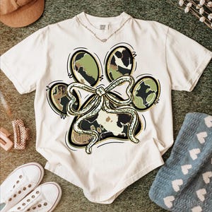 K&ouml;nnte beinhalten: Cremefarbenes T-Shirt mit einem Pfotenabdruck-Design mit einer Schleife. Der Pfotenabdruck hat ein Camouflage-Muster mit goldenen Akzenten. Das Shirt ist von einem braunen Hut, wei&szlig;en Turnschuhen und einer blauen Socke umgeben.