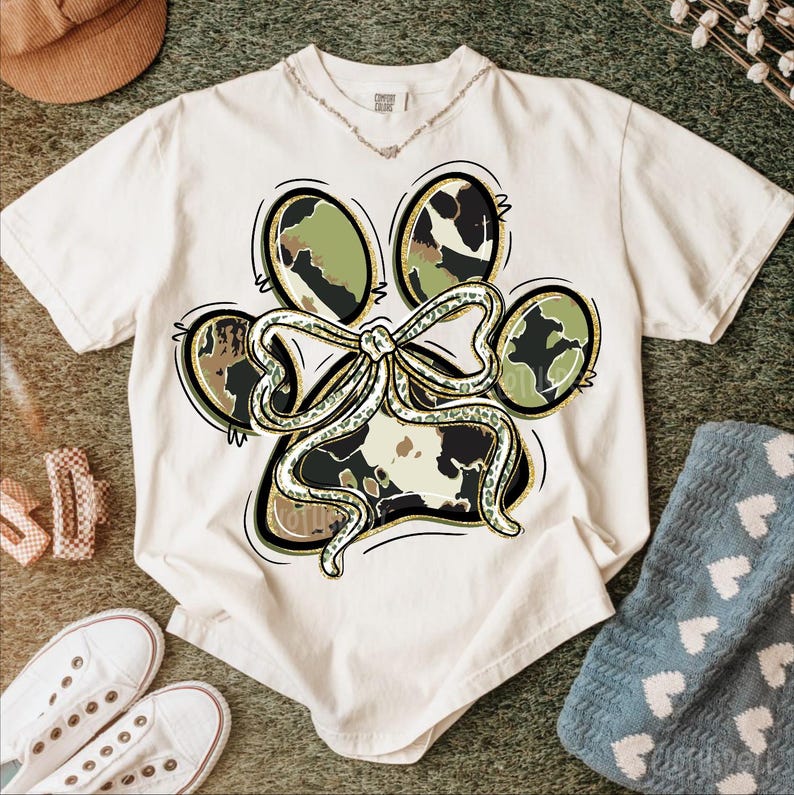 K&ouml;nnte beinhalten: Cremefarbenes T-Shirt mit einem Pfotenabdruck-Design mit einer Schleife. Der Pfotenabdruck hat ein Camouflage-Muster mit goldenen Akzenten. Das Shirt ist von einem braunen Hut, wei&szlig;en Turnschuhen und einer blauen Socke umgeben.