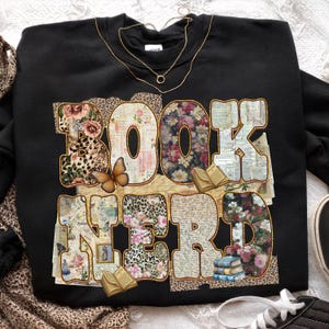 May include: Black sweatshirt with the words "BOOK NERD" in a large, decorative font. The letters are filled with floral, leopard print, and vintage paper patterns. A butterfly and small books are also featured.