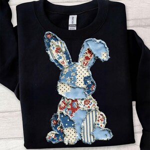 May include: Black sweatshirt featuring a patchwork bunny design. The bunny is made of various fabrics, including floral, polka dot, and striped patterns in red, blue, and white. The sweatshirt has a crew neck and long sleeves.