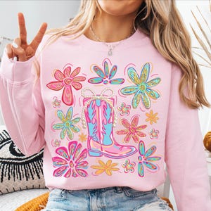 May include: Pink sweatshirt featuring a floral design and a cowboy boot with a pearl necklace. The flowers are in shades of pink, blue, and yellow with gold glitter accents. The boot is pink and blue. A fun and colorful design.