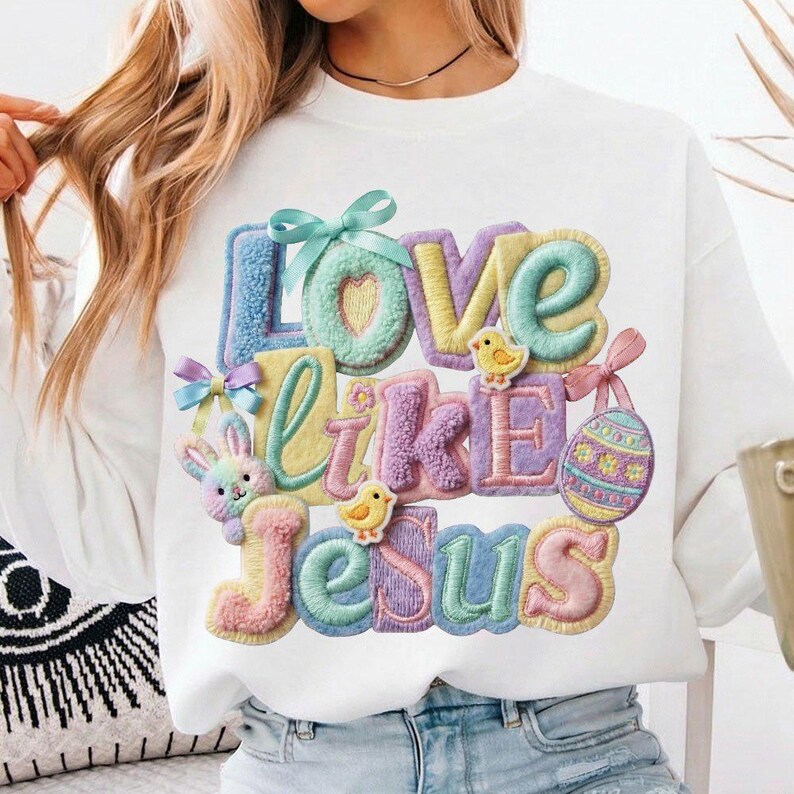 Pu&ograve; includere: Felpa bianca con la scritta "Love Like Jesus" in lettere colorate e strutturate. Il design include un cuore, un coniglietto, pulcini e un uovo di Pasqua, il tutto in colori pastello. La felpa ha uno scollo tondo.