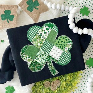 Puede incluir: Camiseta negra de manga larga con un gran dise&ntilde;o de tr&eacute;bol de retazos en varios tonos de verde y blanco. La camiseta est&aacute; decorada con un tr&eacute;bol y monedas de oro, perfecta para el D&iacute;a de San Patricio.