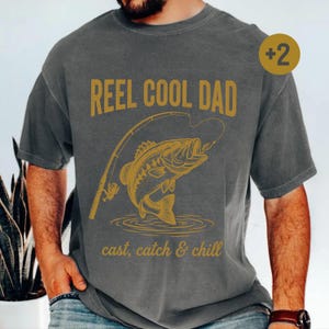 May include: Grey t-shirt featuring a gold illustration of a fish and fishing rod, with the text "REEL COOL DAD" and "cast, catch & chill". The shirt is a casual style and suitable for everyday wear.