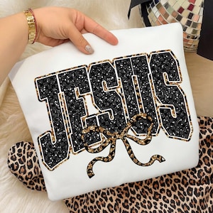 May include: White sweatshirt with the word "JESUS" in a black and gold glitter design. The letters have a black and white checkered pattern. A gold bow is below the word. The sweatshirt is displayed on a leopard print background.