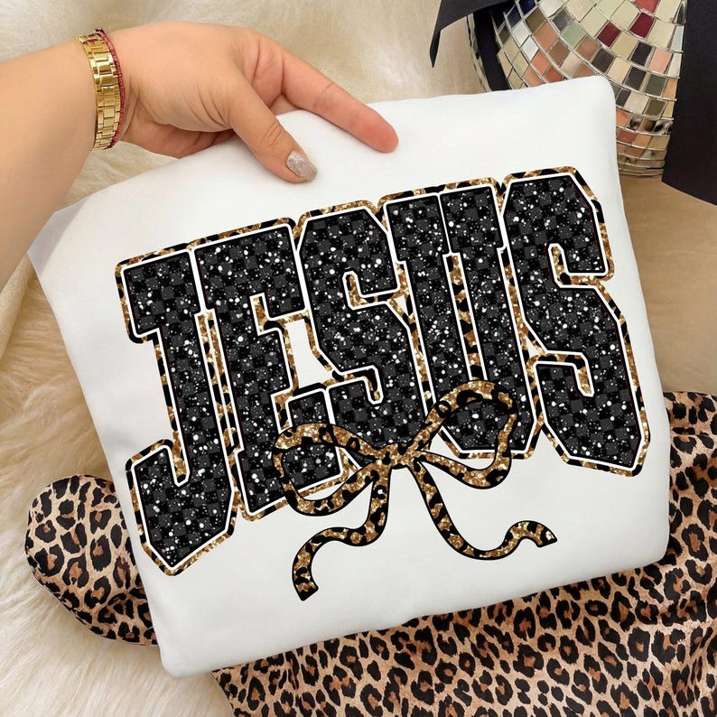 May include: White sweatshirt with the word "JESUS" in a black and gold glitter design. The letters have a black and white checkered pattern. A gold bow is below the word. The sweatshirt is displayed on a leopard print background.