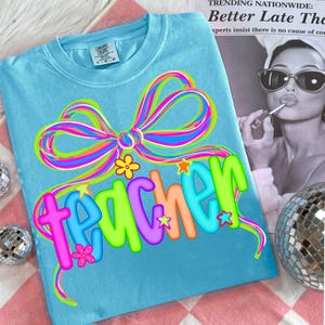 May include: A light blue Comfort Colors t-shirt with a colorful bow and the word "teacher" in a playful font. The design includes flowers and stars. The shirt is a comfortable, casual style.