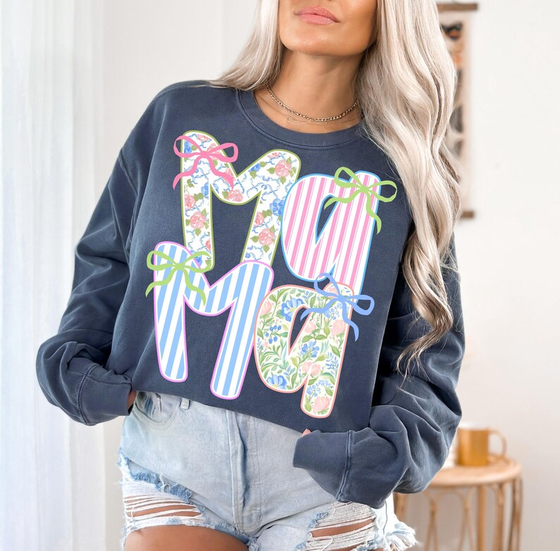 May include: A navy blue sweatshirt with the word "Ma" in a floral pattern and pink stripes, accented with bows. The sweatshirt is a relaxed fit, perfect for casual wear.