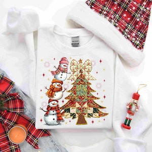 May include: White sweatshirt featuring a Christmas tree and three snowmen wearing scarves and hats. The tree is decorated with a bow and star accents. A Santa hat and a nutcracker ornament are also visible.