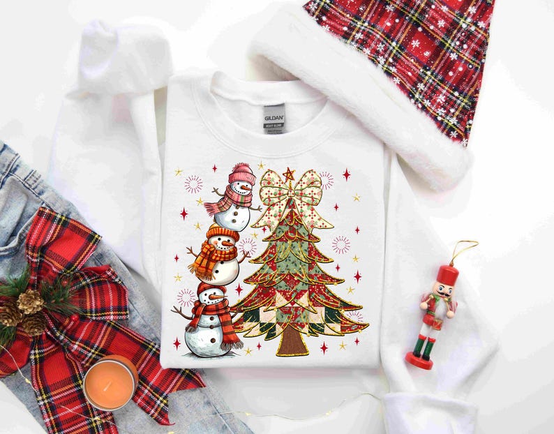 May include: White sweatshirt featuring a Christmas tree and three snowmen wearing scarves and hats. The tree is decorated with a bow and star accents. A Santa hat and a nutcracker ornament are also visible.