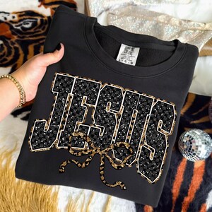 May include: Black sweatshirt with the word "JESUS" in a bold, glittery design. The letters are outlined in white and filled with a black and white checkered pattern and gold glitter. A gold leopard print bow is below the word.