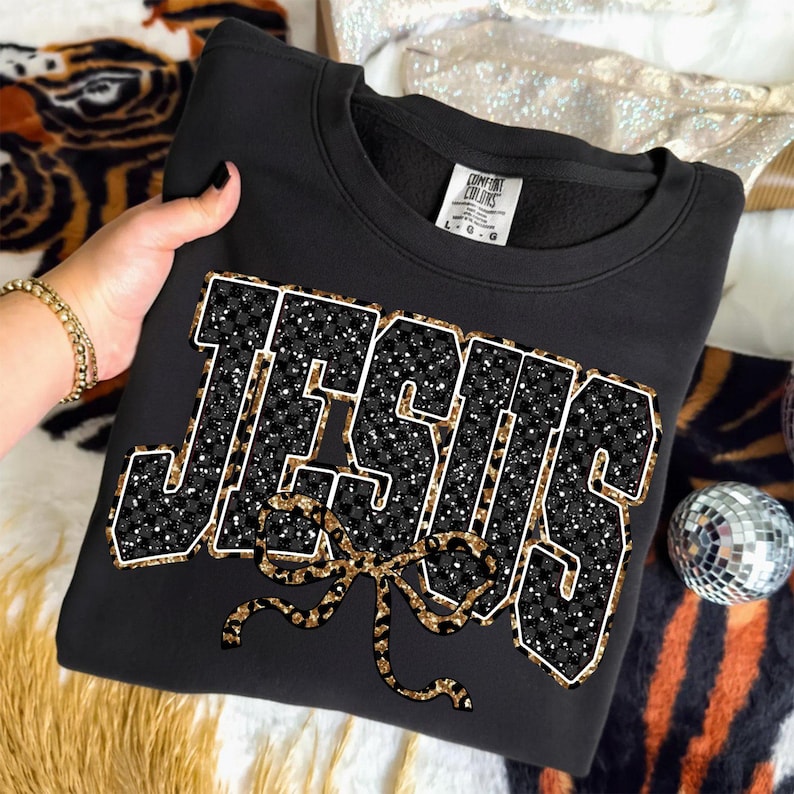 May include: Black sweatshirt with the word "JESUS" in a bold, glittery design. The letters are outlined in white and filled with a black and white checkered pattern and gold glitter. A gold leopard print bow is below the word.
