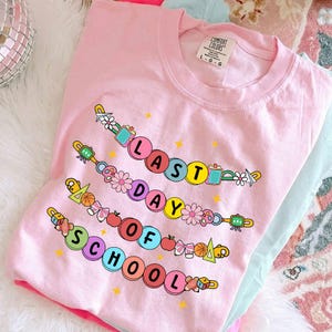 May include: Pink Comfort Colors t-shirt with the text "LAST DAY OF SCHOOL" in colorful, circular letters. The design includes school-themed graphics like apples, flowers, and pencils. The shirt is a size L-G-G.