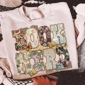 May include: A cream-colored sweatshirt with the words "BOOK NERD" in a collage of floral, leopard print, and vintage paper patterns. A butterfly and small book illustrations are also featured in the design.