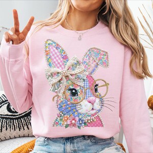 May include: A light pink sweatshirt featuring a colorful patchwork bunny design. The bunny has glasses, a bow, and pearl accents. The bunny's ears are decorated with a gingham pattern. The sweatshirt is a comfortable, casual style.