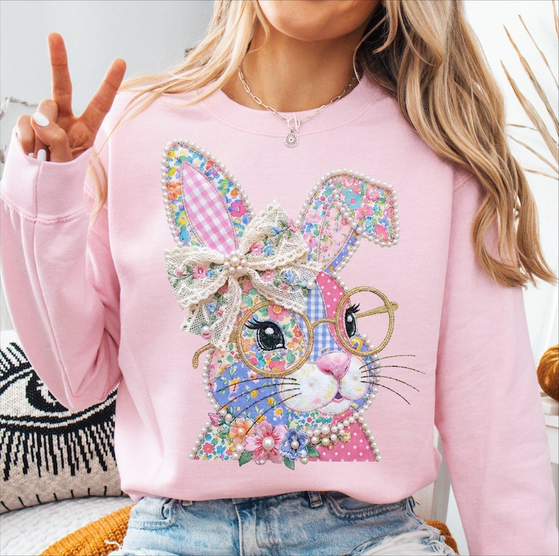 May include: A light pink sweatshirt featuring a colorful patchwork bunny design. The bunny has glasses, a bow, and pearl accents. The bunny's ears are decorated with a gingham pattern. The sweatshirt is a comfortable, casual style.