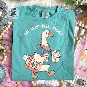 May include: A teal t-shirt featuring a graphic of a goose dressed as a nurse, wearing a patterned neckerchief, carrying a medical bag with a blue star symbol, holding a coffee cup and a clipboard, with the text "OFF TO DO NURSE THINGS" above.