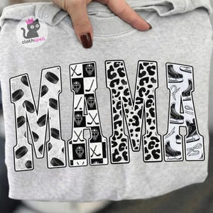 May include: A light grey sweatshirt featuring the word MAMA in large block letters. Each letter has a unique pattern: hockey pucks, a checkerboard with goalie masks and sticks, a black and white leopard print, and ice hockey skates.