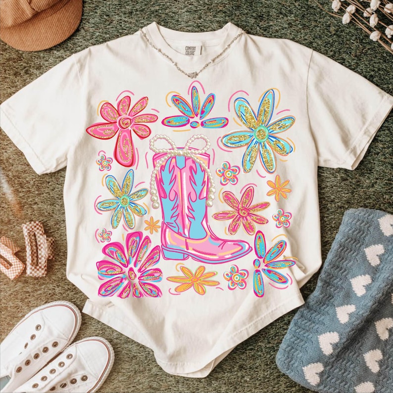 May include: White t-shirt with a colorful floral and cowboy boot design. The graphic features pink, blue, and yellow flowers surrounding a pink and blue cowboy boot with a pearl necklace. A silver necklace is visible at the neckline.