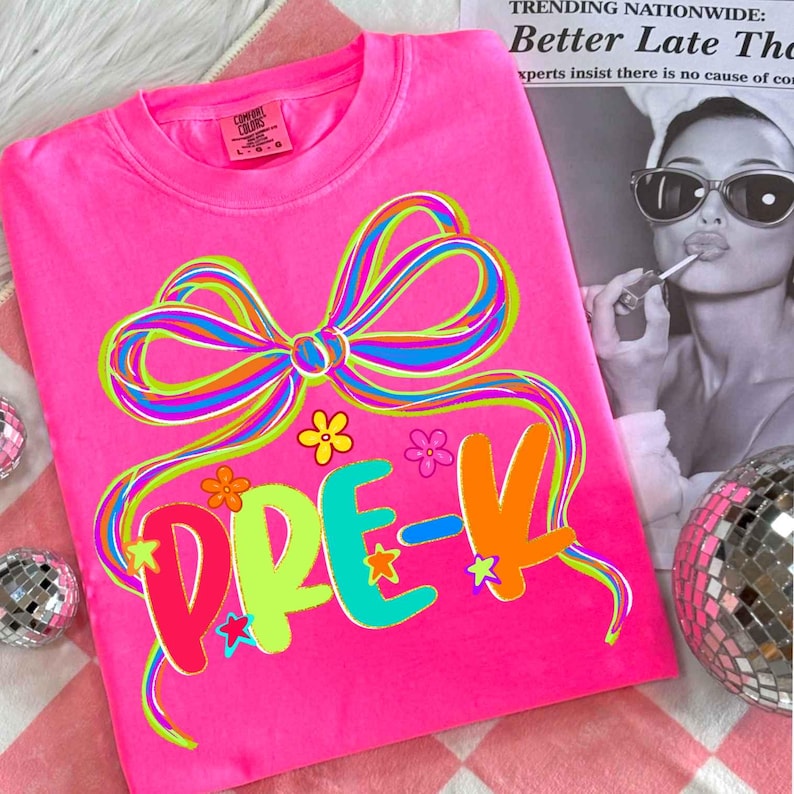May include: Bright pink long-sleeved t-shirt with a colorful bow and the word "PRE-K" in vibrant letters. The design includes flowers and stars. The shirt is made of a soft material, perfect for preschool.
