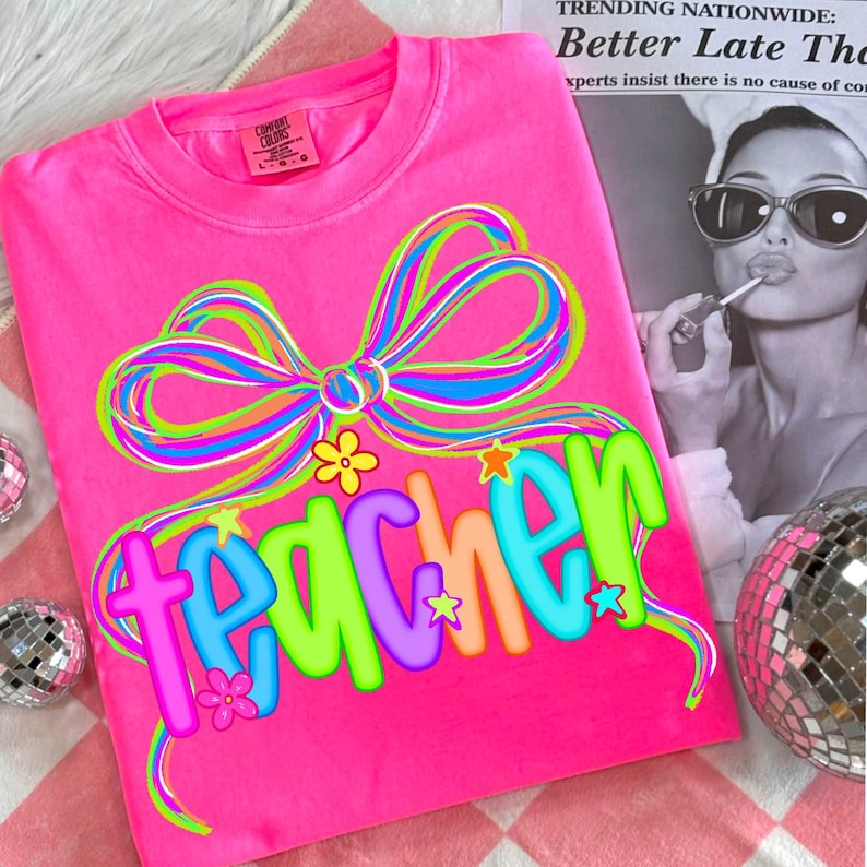 May include: A bright pink t-shirt with the word "teacher" in colorful, bubble letters. A large, multi-colored bow graphic sits above the word. The shirt is a Comfort Colors brand.