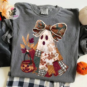 Puede incluir: Una camiseta gris carbón con un gráfico de un fantasma hecho de varios parches de tela, con un gran lazo floral. El fantasma sostiene un cubo de calabaza, con tallos de trigo y hojas de otoño situados a la izquierda de la figura.