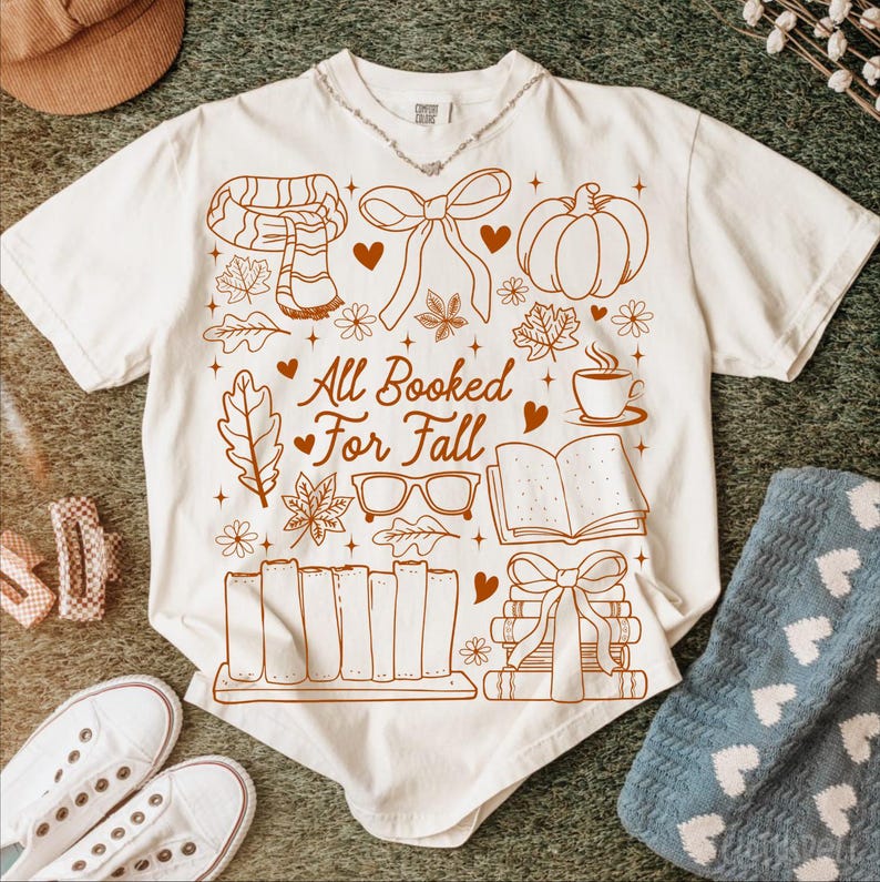 May include: Cream-colored t-shirt with a brown graphic design featuring fall-themed illustrations, including a scarf, pumpkin, leaves, and books. The text "All Booked For Fall" is also included in the design. The shirt is displayed on a green surface.
