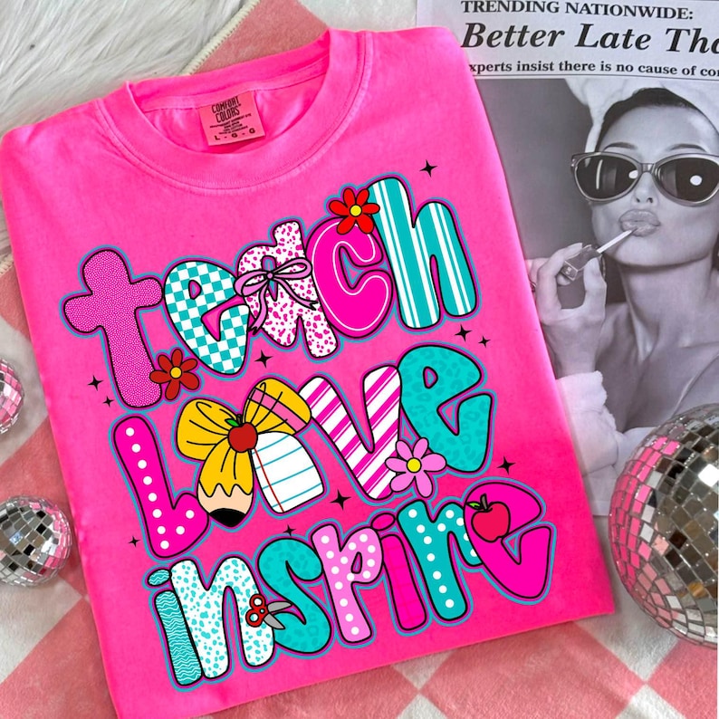 May include: A bright pink Comfort Colors sweatshirt with the words "Teach Love Inspire" in a colorful, playful font. The design includes flowers, bows, and an apple. The shirt is a crew neck and has a relaxed fit.
