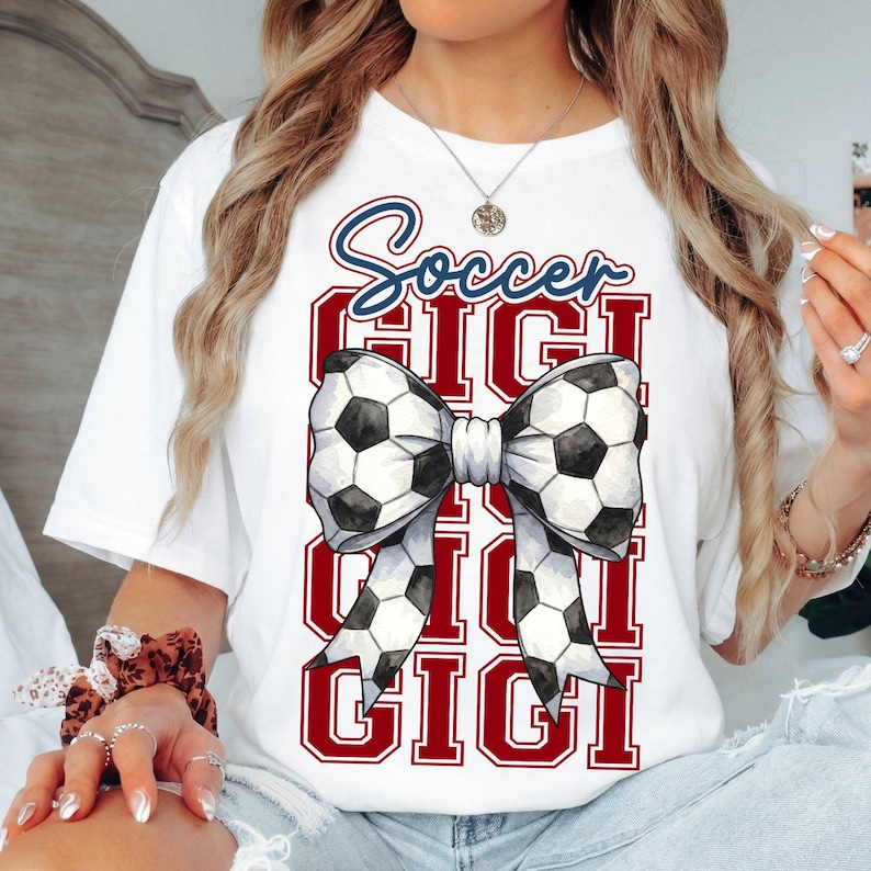 May include: White t-shirt with a soccer-themed design. The design features the word "Soccer" in blue script above a bow made of soccer balls. Below the bow, the word "GIGI" is repeated in a red, block-letter style.