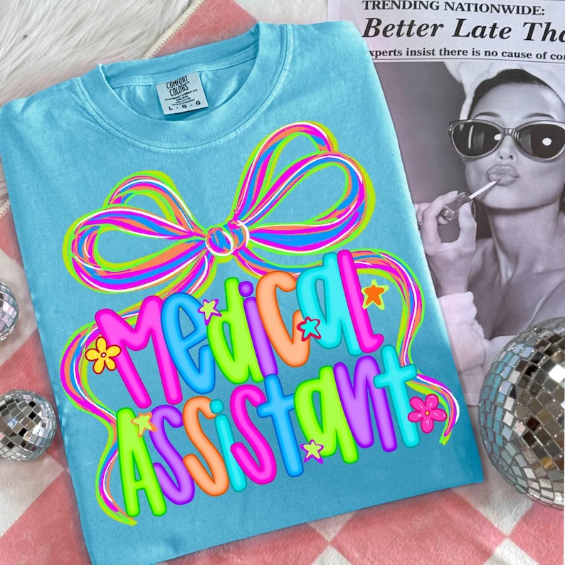 Puede incluir: Camiseta azul claro con la frase "Medical Assistant" en letras coloridas. El dise&ntilde;o incluye un gran lazo multicolor y detalles de estrellas y flores. La camiseta est&aacute; hecha de un material suave.