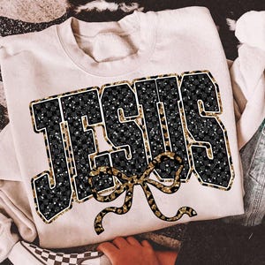May include: Cream-colored sweatshirt with the word "JESUS" in large, bold letters. The letters are black with a checkered pattern and gold glitter accents. A gold bow is below the word. The sweatshirt is casual wear.