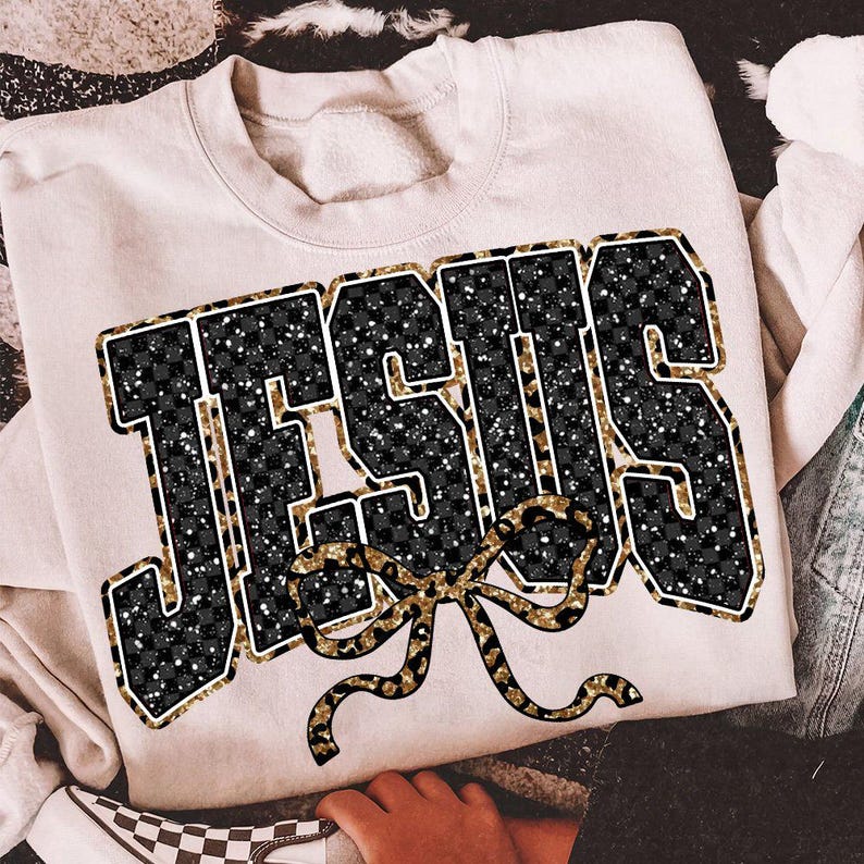 May include: Cream-colored sweatshirt with the word "JESUS" in large, bold letters. The letters are black with a checkered pattern and gold glitter accents. A gold bow is below the word. The sweatshirt is casual wear.