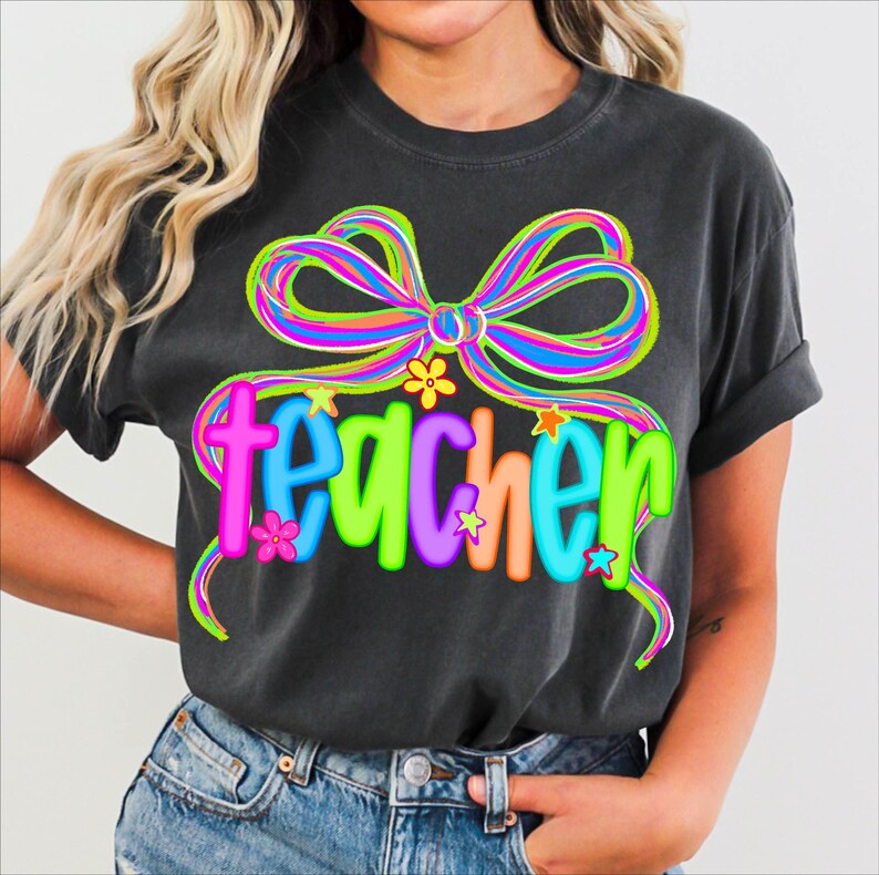 May include: Dark gray t-shirt with a colorful bow and the word "teacher" in bright, playful letters. The design includes flowers and stars, creating a cheerful and vibrant look. A comfortable tee for teachers.