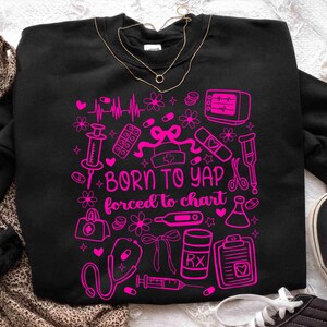 May include: Black sweatshirt featuring a pink medical graphic with a stethoscope, syringe, and pills. The text reads "BORN TO YAP forced to chart". A gold necklace is draped on the sweatshirt.