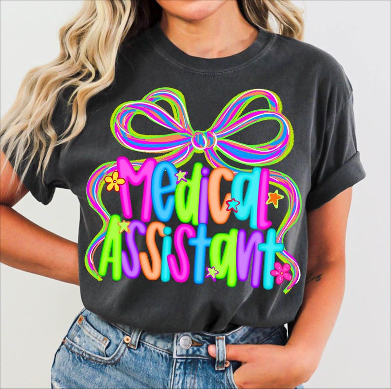 Puede incluir: Camiseta gris oscuro con la frase "Medical Assistant" en un dise&ntilde;o colorido y divertido. El dise&ntilde;o incluye un gran lazo multicolor, flores y estrellas. La camiseta es de estilo casual.