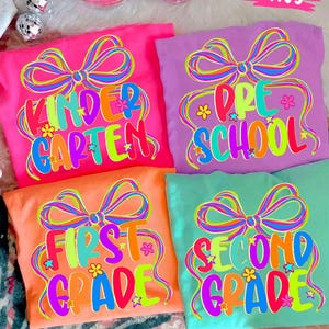 May include: Four colorful t-shirts with grade level text: Kindergarten, Pre-School, First Grade, and Second Grade. Each shirt features a large bow design and the grade level in bright, multi-colored text. The shirts are pink, purple, orange, and teal.