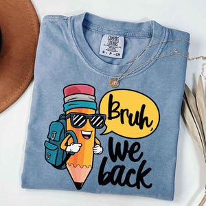 May include: A light blue t-shirt featuring a cartoon pencil character wearing sunglasses and a backpack. The pencil is smiling and has a speech bubble that says "Bruh we back" in black lettering. A gold necklace is draped across the shirt.