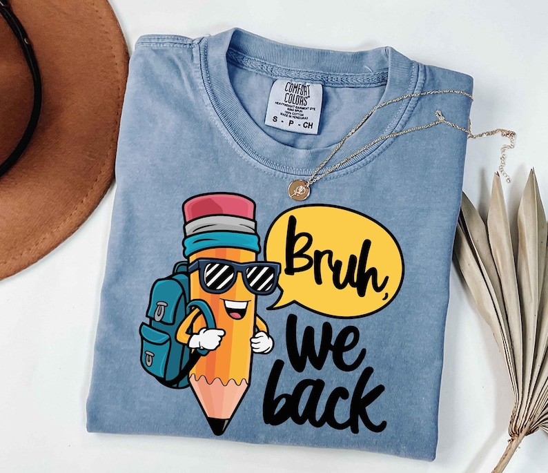 May include: A light blue t-shirt featuring a cartoon pencil character wearing sunglasses and a backpack. The pencil is smiling and has a speech bubble that says "Bruh we back" in black lettering. A gold necklace is draped across the shirt.