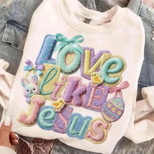 Pu&ograve; includere: Felpa bianca con la frase "Love Like Jesus" in lettere colorate e strutturate. Il design include un coniglietto, pulcini e un uovo di Pasqua, il tutto in colori pastello. Un fiocco azzurro chiaro &egrave; in cima alla parola "Love".