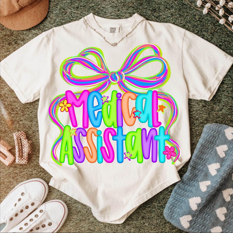 Puede incluir: Camiseta blanca con un gr&aacute;fico colorido que dice "Medical Assistant". El dise&ntilde;o presenta un gran lazo con rayas de arco&iacute;ris y las palabras en colores brillantes. Peque&ntilde;as flores y estrellas acent&uacute;an el dise&ntilde;o. La camiseta est&aacute; sobre un fondo neutro.