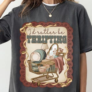 May include: Dark gray t-shirt with a vintage-style graphic. The graphic features a shopping cart filled with dishes, a lamp, and a mirror, with the text "I'd rather be thrifting" in a retro font.