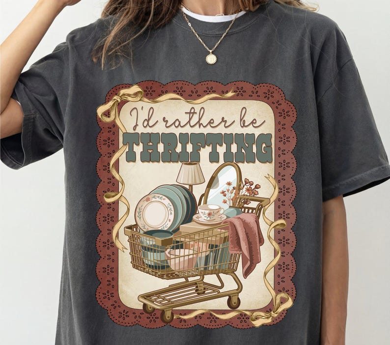 May include: Dark gray t-shirt with a vintage-style graphic. The graphic features a shopping cart filled with dishes, a lamp, and a mirror, with the text "I'd rather be thrifting" in a retro font.