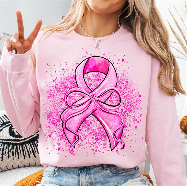 May include: Pink sweatshirt featuring a large pink ribbon design with a bow and a splatter effect. The ribbon has a textured pattern. The sweatshirt is a light pink color.