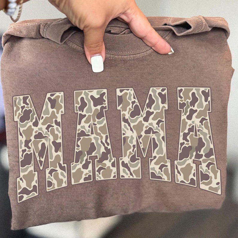 May include: A brown sweatshirt with the word "MAMA" in large, camouflage-patterned letters. The letters are filled with shades of brown, tan, and cream. The sweatshirt is held up by a hand.