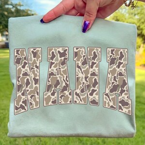 May include: A light green sweatshirt with the word "NANA" in large, block letters. The letters have a camouflage pattern in shades of brown, tan, and gray. The sweatshirt is held up by a hand with purple nail polish.