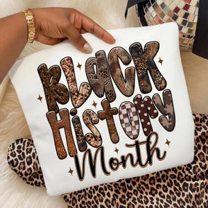 May include: White sweatshirt with the text "BLACK HISTORY MONTH" in a mix of animal print and brown lettering. The sweatshirt is held up by a hand with a gold bracelet. The background includes a leopard print fabric.
